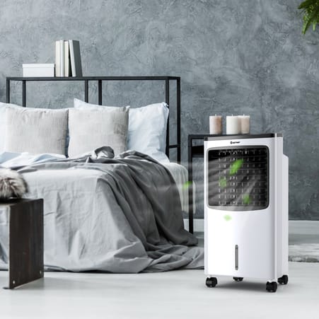 A portable air cooler next to a bed with gray bedding, beside a black metal bedside table with books and candles.