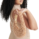 A woman with curly hair holds a beaded handbag with a shimmering pearl-like texture, wearing a beige sleeveless top.