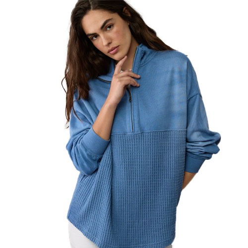 Blue oversized half-zip sweatshirt featuring a textured lower panel and a high collar, worn by a woman with dark hair.