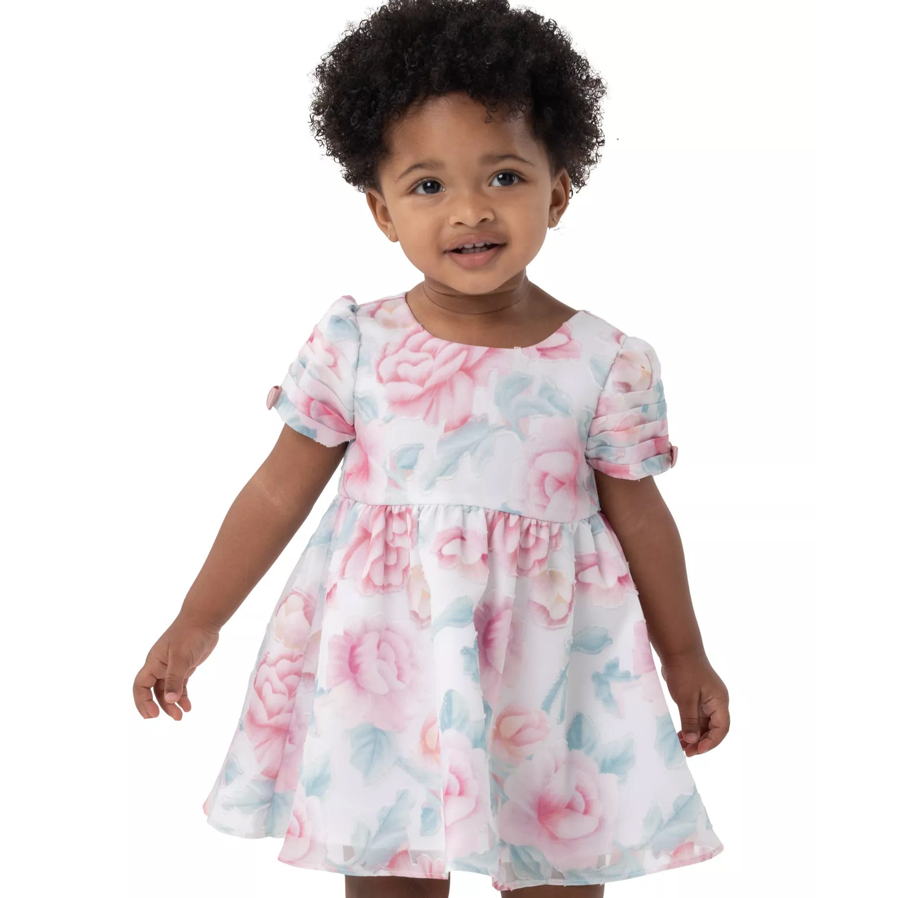 A toddler is wearing a floral dress with short puffed sleeves, featuring pink roses and green leaves on a white background.