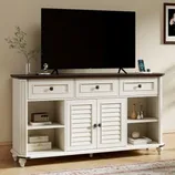 A white TV stand with a dark top features three drawers, two shutter-style doors, and open shelves, displaying decorative items and electronics.
