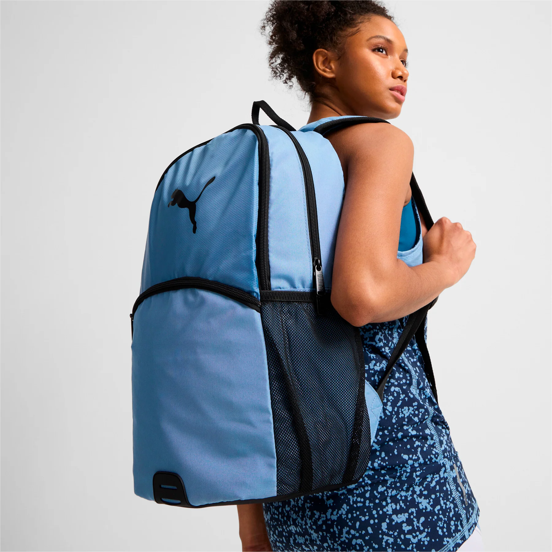 A person wearing a blue tank top is carrying a blue backpack with a visible logo and mesh side pockets.