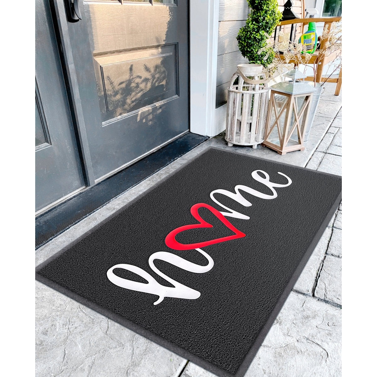 A doormat with the word \“home\“ in white script, with the \“o\“ replaced by a red heart shape, placed in front of a door with decorative lanterns and plants nearby.