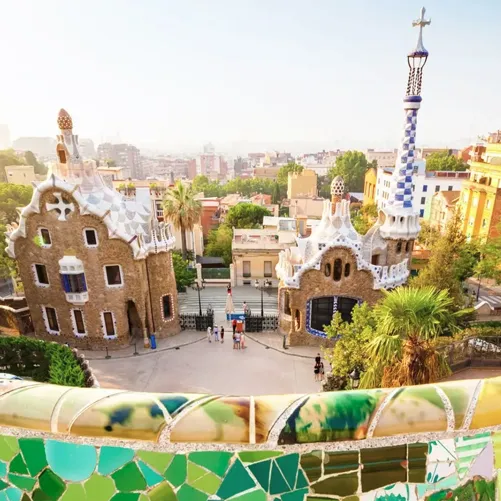 A panoramic view of Park Güell in Barcelona features two whimsical Gaudí-designed buildings with organic shapes and vibrant mosaics set amidst a cityscape.