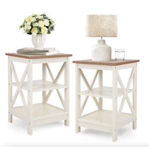 Two white and brown end tables with a cross design on the sides, one with a vase of flowers and one with a lamp and glass.