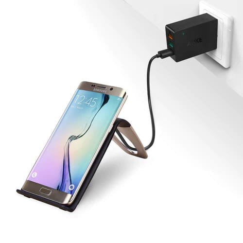 A smartphone is connected to a wall charger with a cable, resting on a mount attached to the outlet.