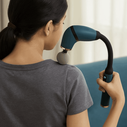 A person is using a handheld massage gun on their shoulder, with a blue handle and a spherical attachment, sitting on a blue couch.