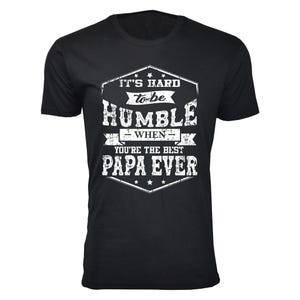 Black T-shirt with white text in various fonts: \“It's hard to be humble when you're the best Papa ever,\“ surrounded by decorative stars and lines.
