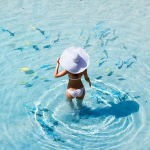 A woman in a white swimsuit and wide-brimmed hat stands in clear blue water surrounded by small fish.