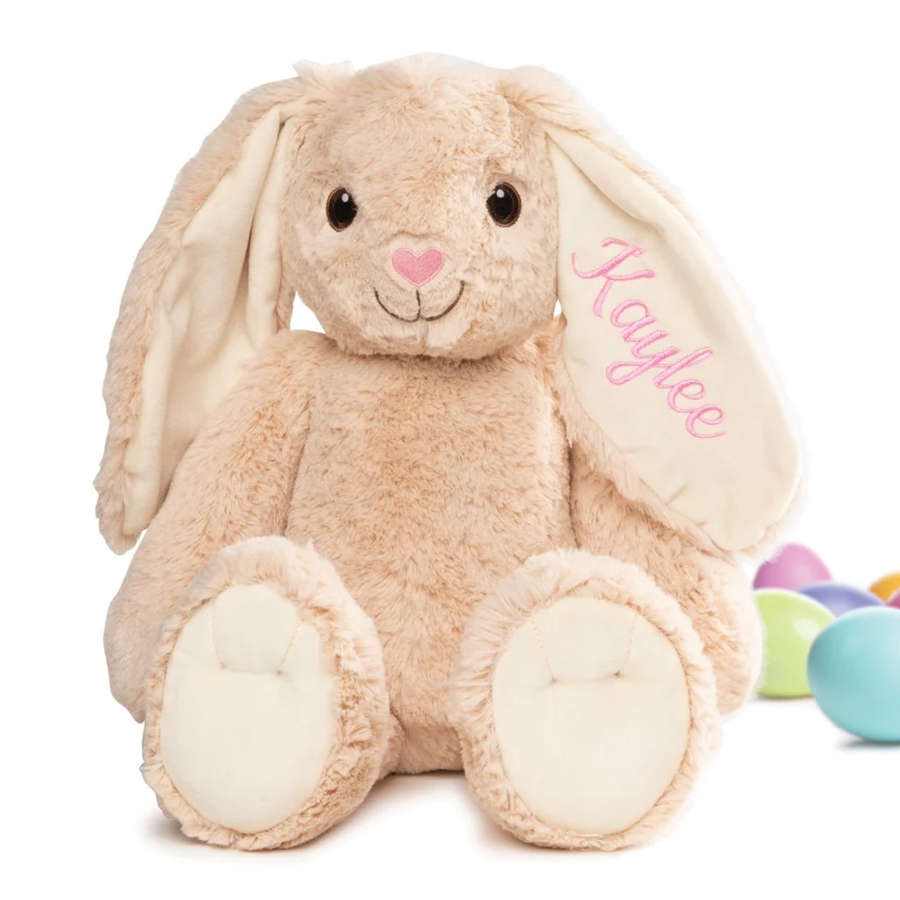 A plush bunny with a beige body, embroidered eyes, and a heart-shaped pink nose, features \“Kaylee\“ embroidered on one ear. Colorful eggs are visible in the background.