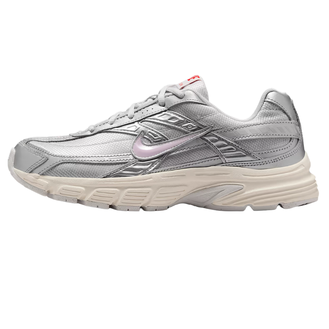 A silver athletic sneaker with a white sole featuring a light pink swoosh logo and red branding on the tongue. The design includes detailed stitching and layered accents.