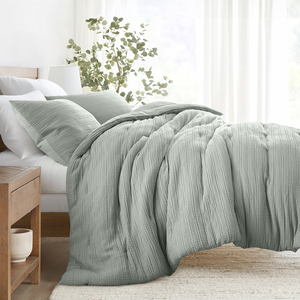 A cozy bed setup featuring a textured, light-colored comforter and matching pillows. A wooden nightstand with a planter is beside the bed, and soft curtains allow diffused light into the room.