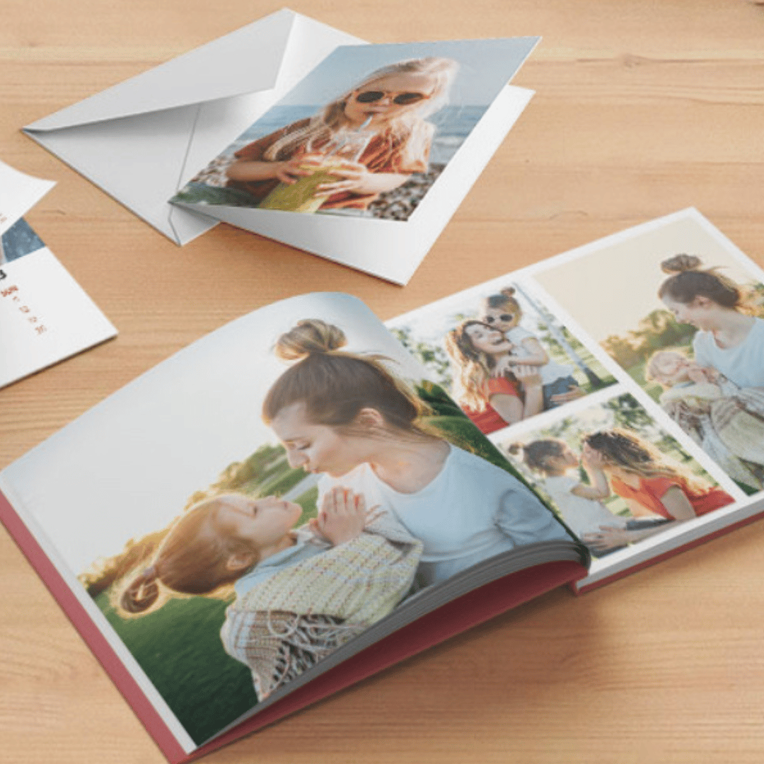 An open photo book and a card with a picture of a girl sipping a drink are displayed on a wooden surface. The book shows images of a woman and child, capturing happy moments together outdoors.