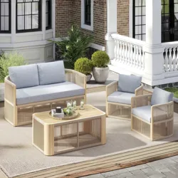 Outdoor patio set with light wood frames and light gray cushions, including a sofa, two chairs, and a matching coffee table on a beige rug, set against a brick and white-paneled house with potted plants.