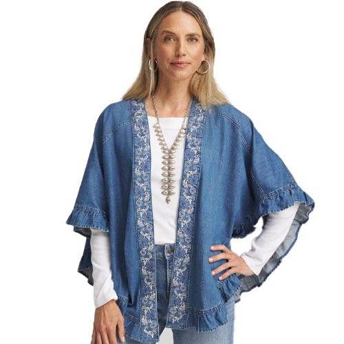 A woman is wearing a blue denim kimono-style jacket with embroidered trim, layered over a white shirt. She accessorizes with a long necklace and hoop earrings.