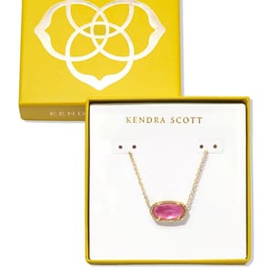 A Kendra Scott necklace with a pink stone on a gold chain is displayed in an open yellow box featuring the brand's logo.