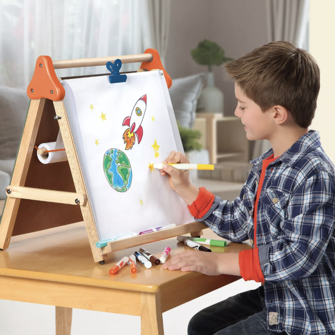 A young boy uses a tabletop easel to draw a colorful space scene, featuring a rocket and the Earth, with markers scattered on the table.
