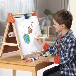 A young boy uses a tabletop easel to draw a colorful space scene, featuring a rocket and the Earth, with markers scattered on the table.