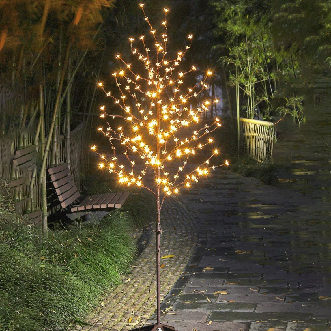 An LED artificial tree light with warm white lights is placed on a garden path next to a wooden bench, creating a decorative outdoor ambiance.