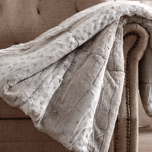 A soft, light gray faux fur throw is draped over the arm of a textured, studded beige chair.