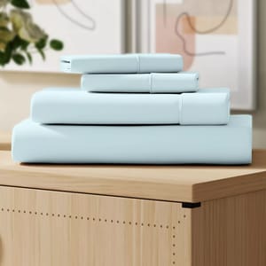 A neatly folded set of light blue bedsheets, placed on a wooden surface, with abstract art in the background.