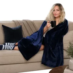 A person sits comfortably on a couch wrapped in a blue fleece blanket, wearing striped socks, and holding a black mug. The setting includes minimalistic decor with a window and a fireplace in the background.