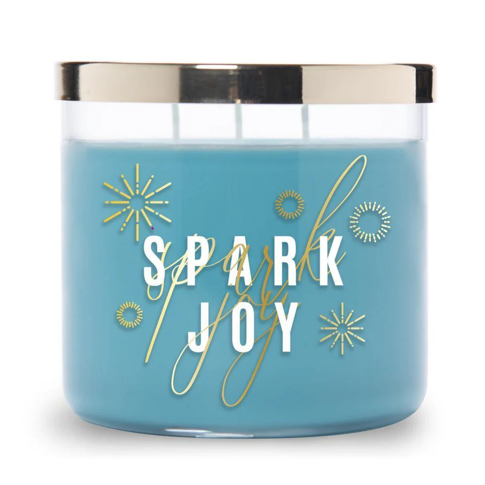 A blue candle with three wicks featuring a metallic lid and the words \“Spark Joy\“ printed in white, accented with decorative gold starburst designs.