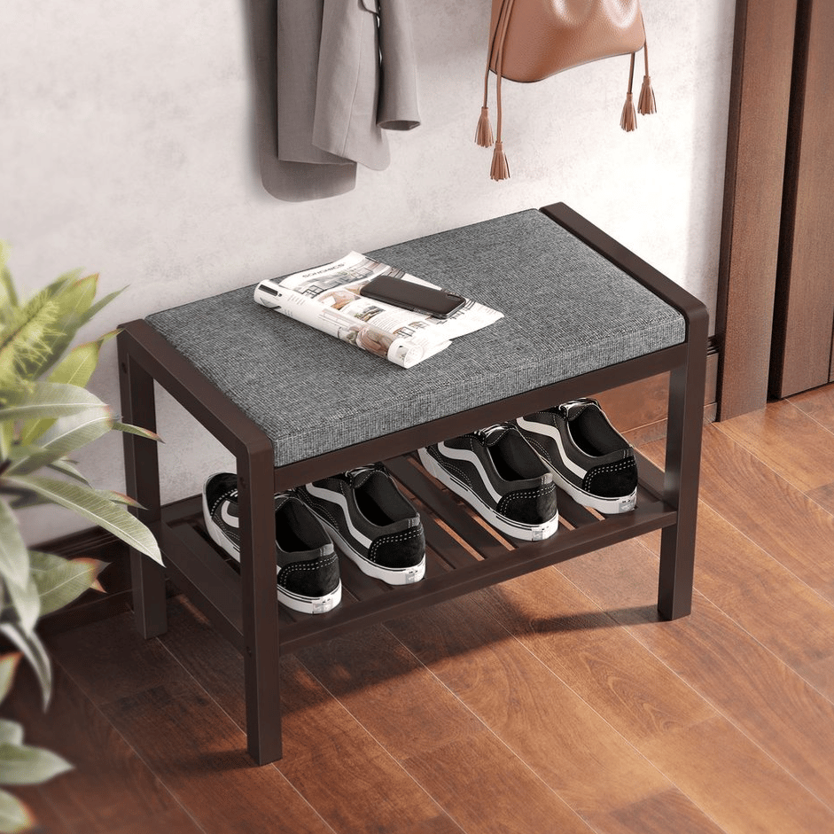 A wooden shoe bench with a gray upholstered seat and a lower slatted shelf holding three pairs of shoes, accompanied by a newspaper and phone on top.