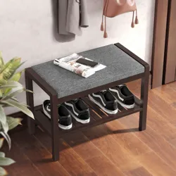 A wooden shoe bench with a gray upholstered seat and a lower slatted shelf holding three pairs of shoes, accompanied by a newspaper and phone on top.