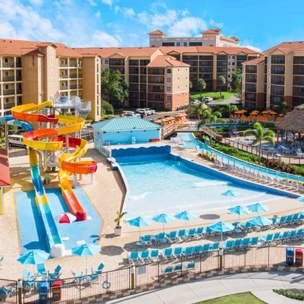 A resort features colorful water slides leading into a pool area with numerous loungers and umbrellas, surrounded by multi-story accommodations and landscaped greenery.