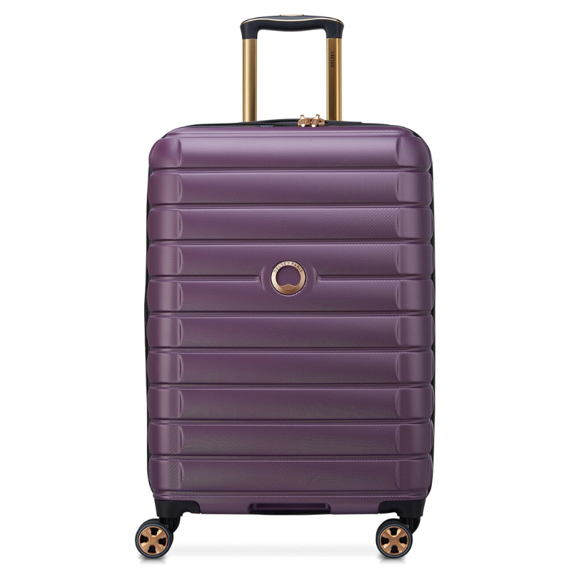 Purple Delsey upright spinner luggage with ribbed texture, telescoping handle, and four dual-spinner wheels. Features a gold accent around the handle and logo.