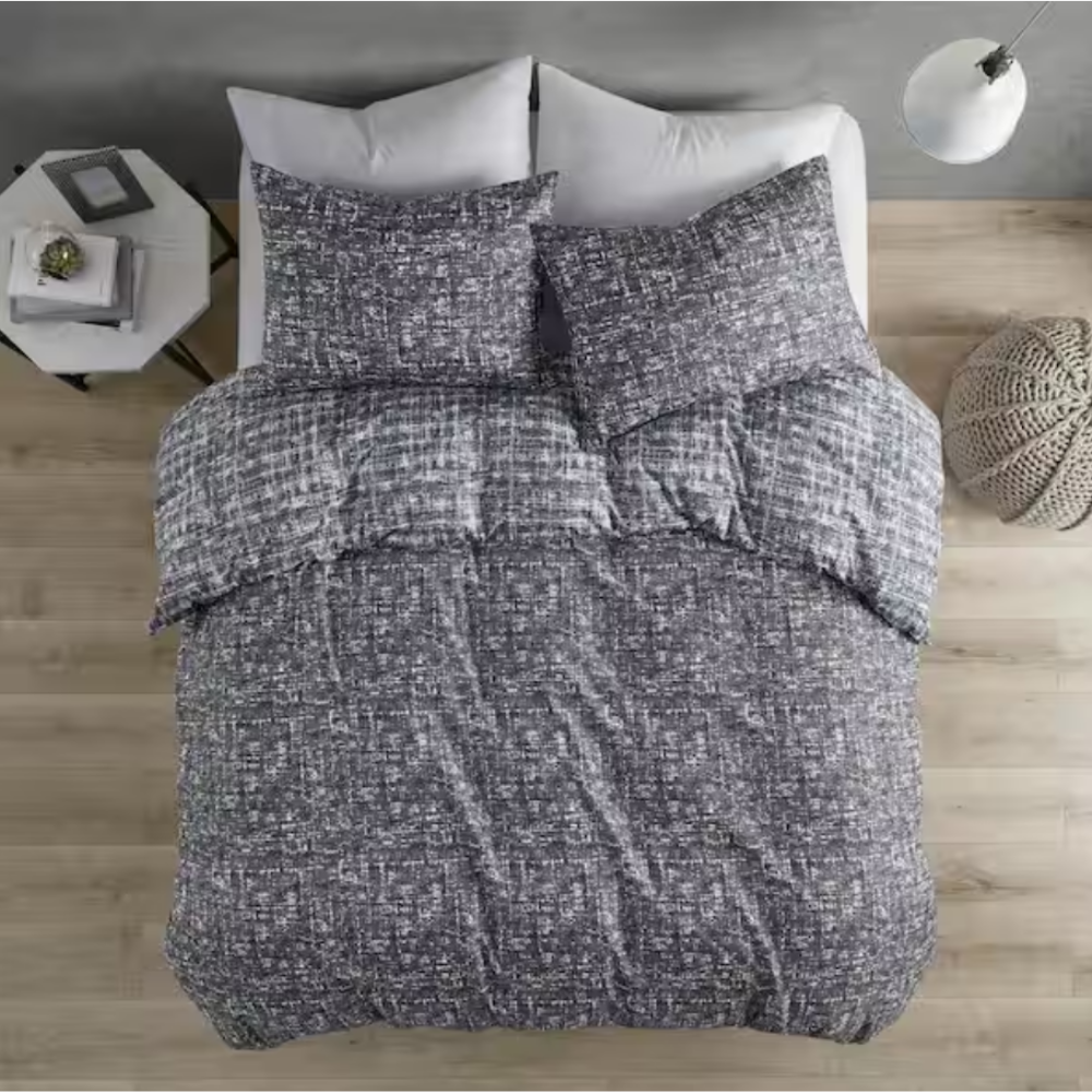 A bed is covered with a textured gray comforter and pillows on a wooden floor, flanked by a side table with books and a knitted pouf. A modern floor lamp stands nearby.
