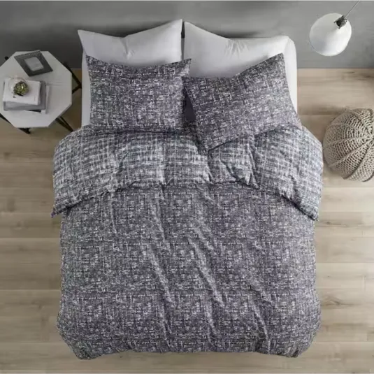 A bed is covered with a textured gray comforter and pillows on a wooden floor, flanked by a side table with books and a knitted pouf. A modern floor lamp stands nearby.