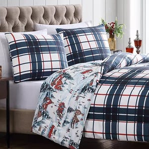 The comforter set features a plaid pattern with blue, red, and white colors on one side and a winter scene with cabins and snowy landscapes on the other. It includes matching pillowcases and is placed on a tufted headboard bed in a bright room.