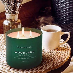 A green Woodland Retreat candle by Colonial Candle is shown, accompanied by a steaming mug on a woven mat. A jar with dried lavender is in the background.