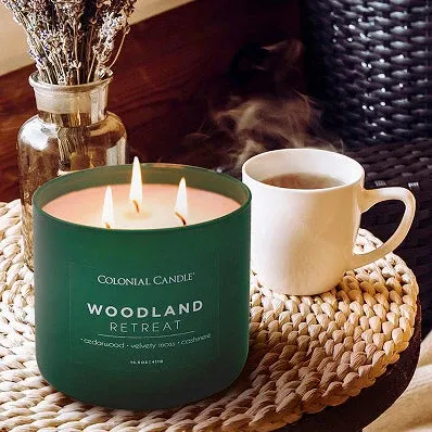 A green Woodland Retreat candle by Colonial Candle is shown, accompanied by a steaming mug on a woven mat. A jar with dried lavender is in the background.