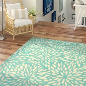 A teal and cream floral-patterned area rug is placed on a hardwood floor beside a rattan armchair with white cushions.