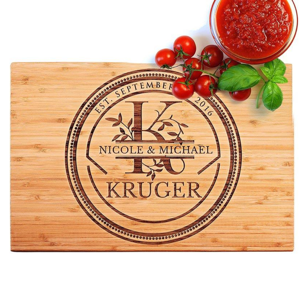 Engraved wooden cutting board featuring \“Nicole & Michael Kruger\“ with a decorative monogram and the date \“Est. September 2016,\“ accompanied by cherry tomatoes, basil, and a bowl of red sauce.