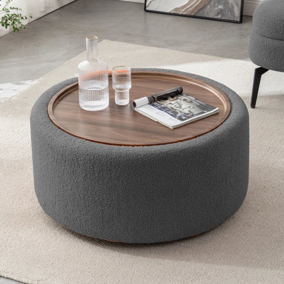 Round ottoman with a wooden tray top, holding a glass carafe, a tumbler, and a magazine. The setting is a modern living room with a white rug, and a gray chair is partially visible.