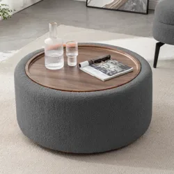 Round ottoman with a wooden tray top, holding a glass carafe, a tumbler, and a magazine. The setting is a modern living room with a white rug, and a gray chair is partially visible.