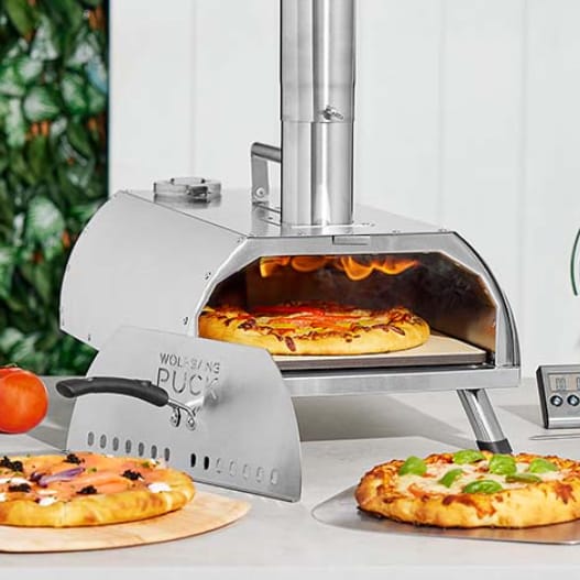 A stainless steel outdoor pizza oven with a pizza cooking inside and another finished pizza with a pizza cutter in front.