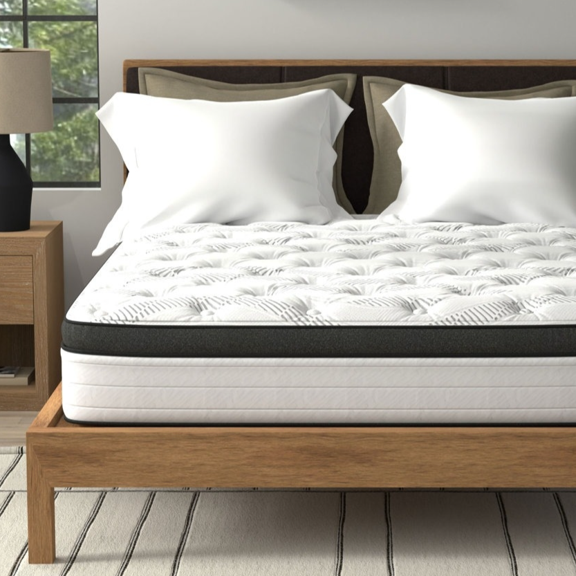 A neatly made bed with a white and black tufted mattress sits on a wooden platform, accompanied by two white pillows. The room includes a nightstand with a lamp, a potted plant, and a framed wall art piece.