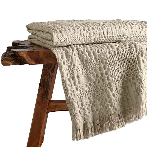A textured cream-colored quilt with a patterned design and fringes on one end is draped over a rustic wooden bench.