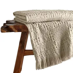 A textured cream-colored quilt with a patterned design and fringes on one end is draped over a rustic wooden bench.