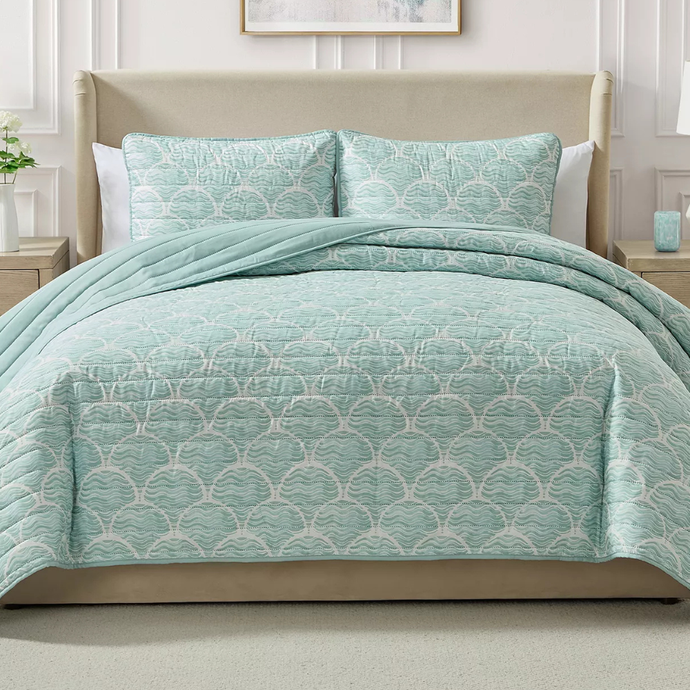 A bed is dressed with a light blue quilt featuring a shell pattern, accompanied by matching shams. The room includes a beige upholstered headboard and light-colored nightstands with a vase and a small decor item.