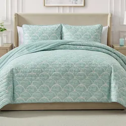 A bed is dressed with a light blue quilt featuring a shell pattern, accompanied by matching shams. The room includes a beige upholstered headboard and light-colored nightstands with a vase and a small decor item.