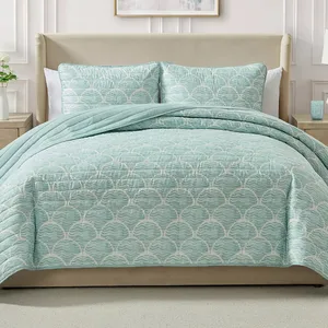 A bed is dressed with a light blue quilt featuring a shell pattern, accompanied by matching shams. The room includes a beige upholstered headboard and light-colored nightstands with a vase and a small decor item.