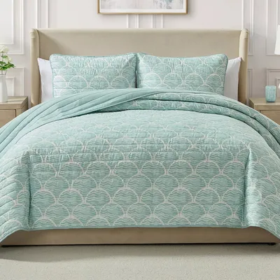 A bed is dressed with a light blue quilt featuring a shell pattern, accompanied by matching shams. The room includes a beige upholstered headboard and light-colored nightstands with a vase and a small decor item.