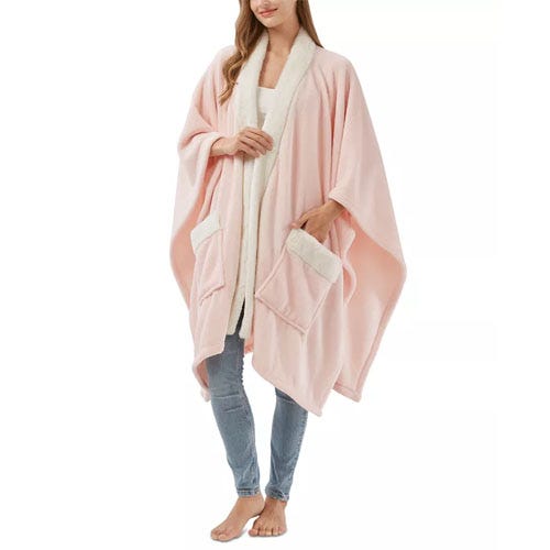 A woman wears a soft pink wearable blanket with a white lining, featuring two front pockets.