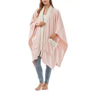 A woman wears a soft pink wearable blanket with a white lining, featuring two front pockets.